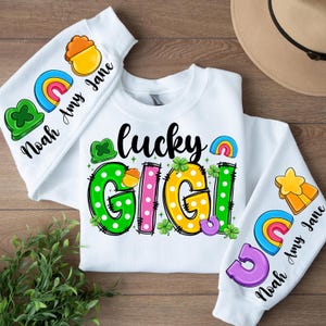 May include: White sweatshirt with the text "lucky gigi" in colorful letters with polka dots and shamrocks. The sleeves have rainbow and pot of gold designs with the names "Noah Amy Jane" on each sleeve.
