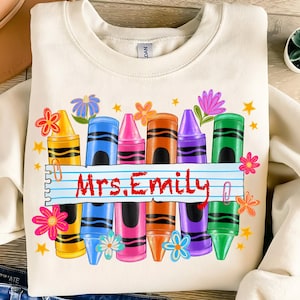 May include: Cream-colored sweatshirt featuring a colorful crayon design. The design includes a row of crayons in various colors, flowers, and stars. The name "Mrs. Emily" is written in red on a lined paper banner.