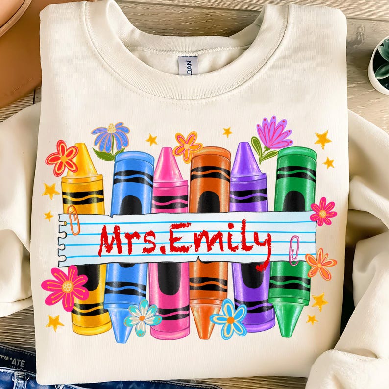 May include: Cream-colored sweatshirt featuring a colorful crayon design. The design includes a row of crayons in various colors, flowers, and stars. The name "Mrs. Emily" is written in red on a lined paper banner.