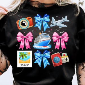 Puede incluir: Camiseta negra con un dise&ntilde;o de tem&aacute;tica de viaje. El dise&ntilde;o presenta un crucero, un avi&oacute;n, una c&aacute;mara, una palmera, un pasaporte y lazos en rosa, azul y blanco.