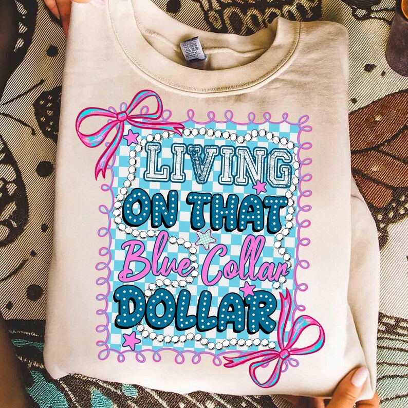 Pu&ograve; includere: Una felpa color crema con la scritta "LIVING ON THAT Blue Collar DOLLAR" in un design blu e rosa. Il design include uno sfondo a scacchi, stelle e fiocchi.