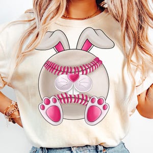 Puede incluir: Una camiseta blanca con un gráfico de béisbol rosa y blanco con la cara de un conejo. El béisbol tiene costuras rosas y una nariz de corazón rosa.