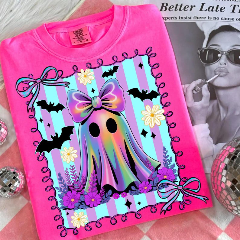 May include: A bright pink Comfort Colors sweatshirt featuring a colorful ghost design with a large bow, bats, flowers, and a striped background. The design is framed with a decorative border. The sweatshirt is a comfortable, casual wear item.