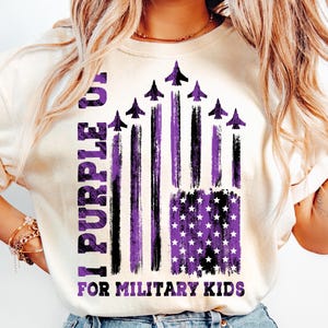 May include: A white t-shirt with a purple and black American flag design. The flag has a distressed look and features purple stars and stripes. The text "I Purple You For Military Kids" is printed on the shirt.