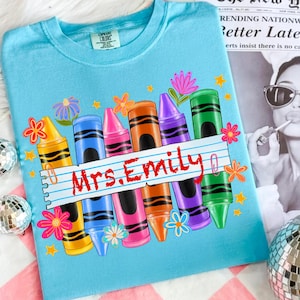 May include: A light blue t-shirt featuring a colorful crayon design with the text "Mrs. Emily" on a lined paper banner. The design includes various colored crayons, flowers, and stars. The shirt is made of a soft material.