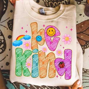 May include: Cream-colored sweatshirt with the words "be kind" in colorful, scribbled letters. The design includes a rainbow, smiley faces, flowers, and a ladybug, creating a cheerful and positive vibe. The sweatshirt is folded.