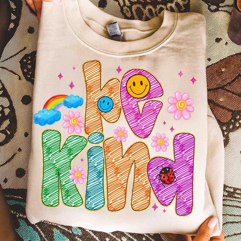 May include: Cream-colored sweatshirt with the words "be kind" in colorful, scribbled letters. The design includes a rainbow, smiley faces, flowers, and a ladybug, creating a cheerful and positive vibe. The sweatshirt is folded.