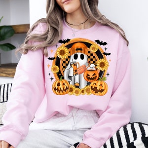 Peut inclure: Sweat-shirt rose clair avec un motif sur le th&egrave;me d'Halloween. Le motif repr&eacute;sente un fant&ocirc;me portant un bonnet orange, tenant une boisson et un seau citrouille. Des tournesols, des chauves-souris et des lanternes entourent le fant&ocirc;me.