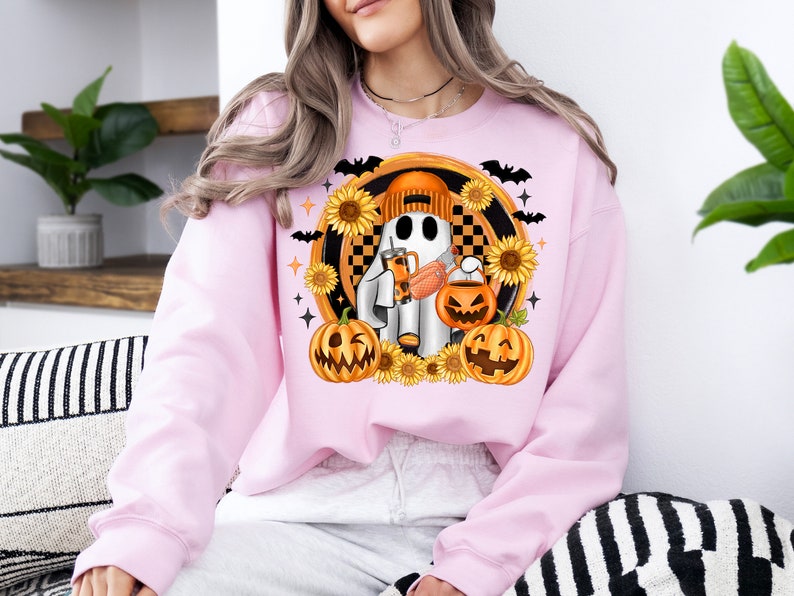 Peut inclure: Sweat-shirt rose clair avec un motif sur le th&egrave;me d'Halloween. Le motif repr&eacute;sente un fant&ocirc;me portant un bonnet orange, tenant une boisson et un seau citrouille. Des tournesols, des chauves-souris et des lanternes entourent le fant&ocirc;me.