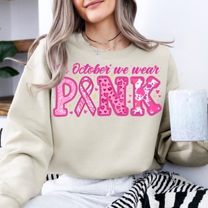 May include: A beige sweatshirt with the text "In October we wear PINK" in pink letters. The letters are decorated with pink hearts, glitter, and a pink ribbon.