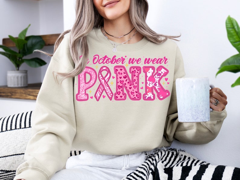 May include: A beige sweatshirt with the text "In October we wear PINK" in pink letters. The letters are decorated with pink hearts, glitter, and a pink ribbon.