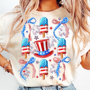Puede incluir: Una camiseta blanca con un dise&ntilde;o rojo, blanco y azul con paletas de hielo, lazos y fuegos artificiales. Las paletas de hielo est&aacute;n decoradas con la bandera estadounidense.