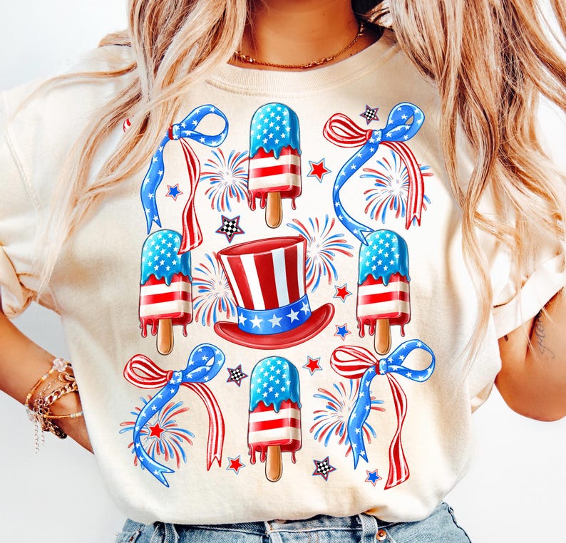 Puede incluir: Una camiseta blanca con un dise&ntilde;o rojo, blanco y azul con paletas de hielo, lazos y fuegos artificiales. Las paletas de hielo est&aacute;n decoradas con la bandera estadounidense.