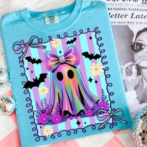 May include: A light blue t-shirt featuring a colorful ghost design. The ghost has a large bow and is surrounded by bats, flowers, and a decorative border. The design includes purple, pink, and blue stripes.