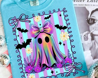 Puede incluir: Una camiseta azul claro con un diseño de fantasma colorido. El fantasma tiene un gran lazo y está rodeado de murciélagos, flores y un borde decorativo. El diseño incluye rayas moradas, rosas y azules.