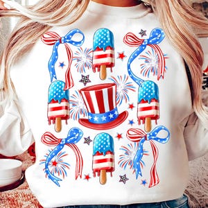 Puede incluir: Sudadera blanca con un tema del 4 de julio en rojo, blanco y azul. El dise&ntilde;o presenta paletas de hielo, lazos, estrellas y fuegos artificiales.