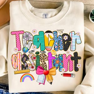 May include: Cream-colored sweatshirt with a colorful graphic that reads "Teacher Assistant." The text is decorated with school-themed elements like pencils, a globe, and a rainbow. The design incorporates various patterns and colors.