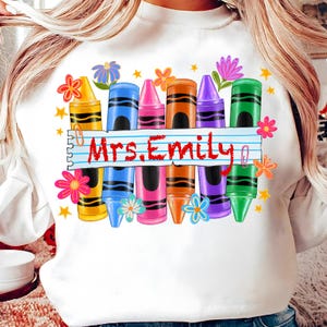 May include: White sweatshirt featuring a colorful crayon design with the text "Mrs. Emily". The design includes yellow, blue, pink, orange, purple, and green crayons, along with flowers and stars.