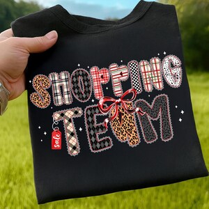 Peut inclure: Sweat-shirt noir avec l'inscription "SHOPPING TEAM" dans diff&eacute;rents motifs, dont l&eacute;opard, carreaux et pois. Un n&oelig;ud rouge et une &eacute;tiquette "sale" sont &eacute;galement pr&eacute;sents.