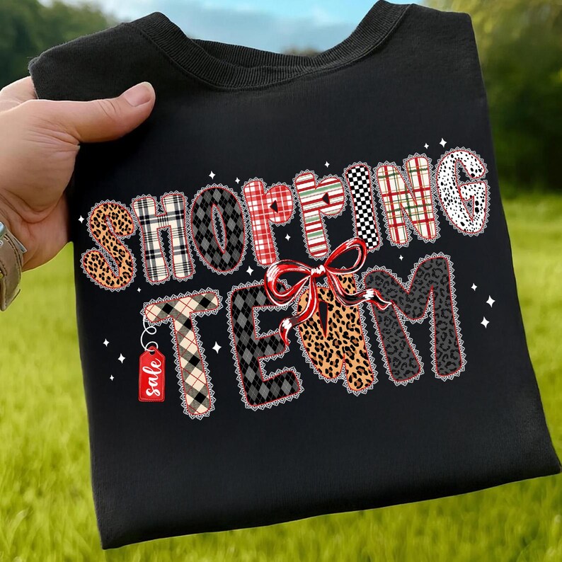 Peut inclure: Sweat-shirt noir avec l'inscription "SHOPPING TEAM" dans diff&eacute;rents motifs, dont l&eacute;opard, carreaux et pois. Un n&oelig;ud rouge et une &eacute;tiquette "sale" sont &eacute;galement pr&eacute;sents.