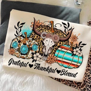 Puede incluir: Coj&iacute;n decorativo color crema con un dise&ntilde;o de calabazas, una calavera de toro y detalles florales. Las calabazas tienen patrones turquesa, de leopardo y a rayas. El texto "Grateful Thankful Blessed" est&aacute; impreso debajo.