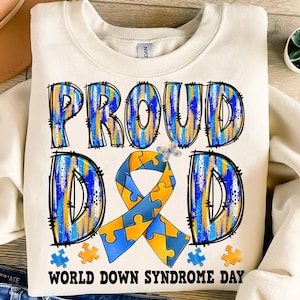 May include: A white sweatshirt with the words "Proud Dad" in blue and yellow puzzle piece patterned lettering. A blue and yellow puzzle piece ribbon is in the center of the shirt. The words "World Down Syndrome Day" are printed below the ribbon.