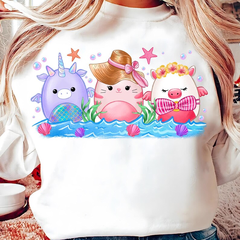 K&ouml;nnte beinhalten: Wei&szlig;er Pullover mit einem Aufdruck von drei Comicfiguren: einem lila Einhorn, einer rosa Katze mit Hut und einem wei&szlig;en Schwein mit einer Blumenkrone. Die Figuren befinden sich in einer Meeresumgebung mit blauen Wellen, Muscheln und Sternen.