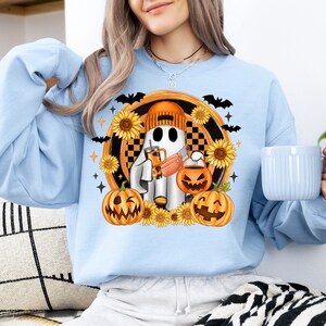 Peut inclure: Sweat-shirt bleu clair avec un motif d'Halloween. Le motif repr&eacute;sente un fant&ocirc;me portant un chapeau, tenant une boisson et une citrouille, entour&eacute; de tournesols, de chauves-souris et de citrouilles. Le sweat-shirt est de style d&eacute;contract&eacute; et confortable.