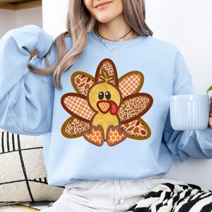 May include: A light blue sweatshirt featuring a patchwork turkey design. The turkey is made from various fabrics, including plaid, leopard print, and honeycomb. The turkey has a yellow body, brown feathers, and a red wattle.