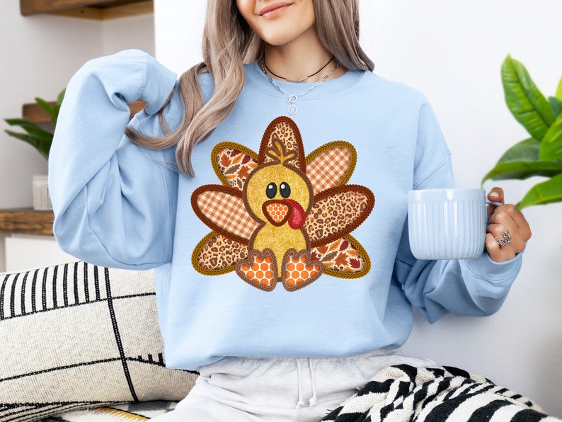 May include: A light blue sweatshirt featuring a patchwork turkey design. The turkey is made from various fabrics, including plaid, leopard print, and honeycomb. The turkey has a yellow body, brown feathers, and a red wattle.