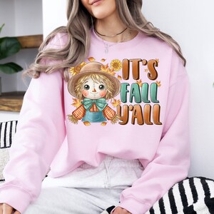 Op de afbeelding: Roze sweatshirt met een cartoon-vogelverschrikker en de tekst "It's Fall Y'all".