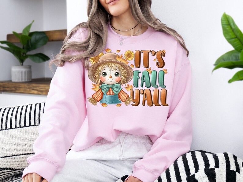 Op de afbeelding: Roze sweatshirt met een cartoon-vogelverschrikker en de tekst "It's Fall Y'all".