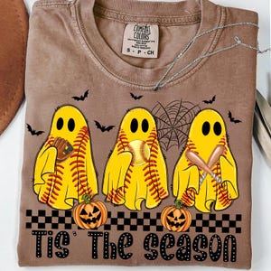 Puede incluir: Camiseta marr&oacute;n con un dise&ntilde;o de Halloween. Tres figuras de fantasmas amarillos est&aacute;n vestidas como pelotas de b&eacute;isbol, bates y un guante. La frase "Tis' The Season" est&aacute; impresa en la parte inferior, junto con dos calabazas y murci&eacute;lagos.