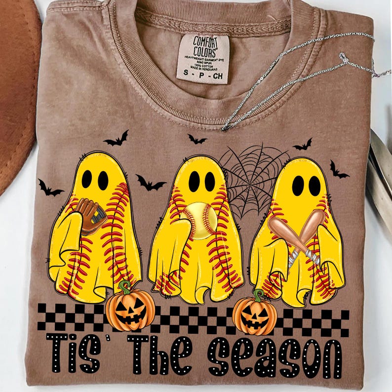 Puede incluir: Camiseta marr&oacute;n con un dise&ntilde;o de Halloween. Tres figuras de fantasmas amarillos est&aacute;n vestidas como pelotas de b&eacute;isbol, bates y un guante. La frase "Tis' The Season" est&aacute; impresa en la parte inferior, junto con dos calabazas y murci&eacute;lagos.