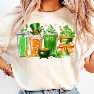 May include: White t-shirt with a graphic of four green, orange, and white drinks with shamrocks, a leprechaun hat, and a pot of gold.