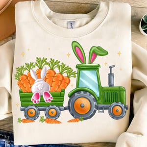 May include: A white sweatshirt with a green tractor pulling a wagon full of carrots. A white bunny rabbit with pink ears is peeking out of the wagon.