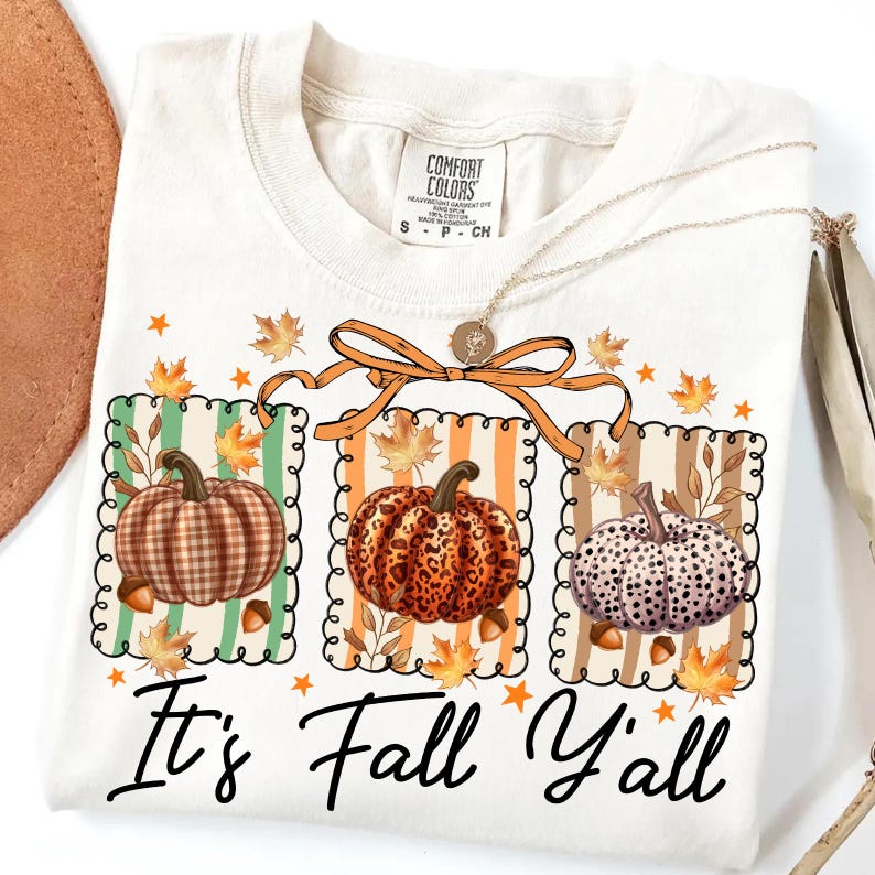 May include: Cream-colored t-shirt with a fall-themed graphic. The graphic features three pumpkins with different patterns, surrounded by leaves and acorns. The text "It's Fall Y'all" is printed below the pumpkins.