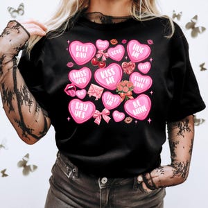 May include: Black t-shirt with pink heart-shaped candy designs. The candy hearts have various messages, including "Best Day", "Kiss Me", "Hug Me", "Sweet Talk", and "Be Mine".