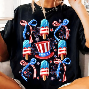 Puede incluir: Camiseta negra con un tema del 4 de julio en rojo, blanco y azul. El dise&ntilde;o presenta paletas de hielo con los colores de la bandera estadounidense, lazos rojos, blancos y azules y fuegos artificiales.