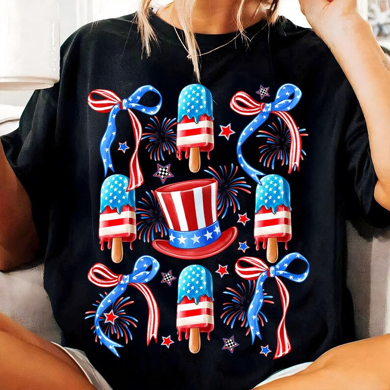 Puede incluir: Camiseta negra con un tema del 4 de julio en rojo, blanco y azul. El dise&ntilde;o presenta paletas de hielo con los colores de la bandera estadounidense, lazos rojos, blancos y azules y fuegos artificiales.