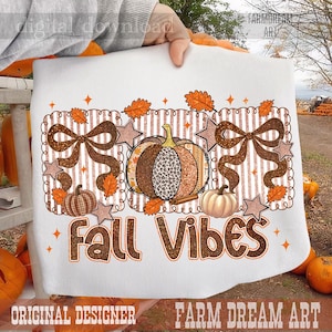 May include: A white t-shirt featuring a graphic design with a central pumpkin, two brown bows, and smaller pumpkins. The design includes orange leaves, stars, and the text Fall Vibes in brown glittery lettering against a striped background.