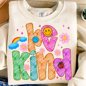 May include: Cream-colored sweatshirt with the words "be kind" in colorful, scribbled letters. The design includes a rainbow, smiley faces, flowers, and a ladybug, creating a cheerful and positive vibe. A cozy and stylish top.