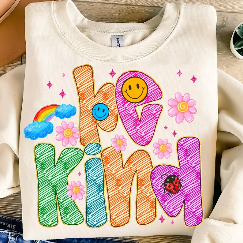 May include: Cream-colored sweatshirt with the words "be kind" in colorful, scribbled letters. The design includes a rainbow, smiley faces, flowers, and a ladybug, creating a cheerful and positive vibe. A cozy and stylish top.