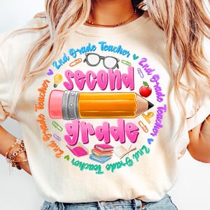 May include: Cream-colored t-shirt with a colorful graphic design for a 2nd-grade teacher. The design features a large pencil, the words "Second Grade" in pink, and other school-related elements like glasses, books, and an apple.