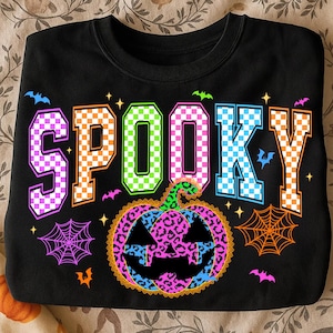 May include: Black sweatshirt with the word "SPOOKY" in colorful checkered letters. A leopard print pumpkin is centered below the word, with bats and spiderwebs. A fun Halloween-themed design.