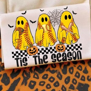 Puede incluir: Camiseta blanca con tres fantasmas amarillos con tem&aacute;tica de softbol, con bates, guantes y una pelota. La frase "Tis' The Season" est&aacute; impresa debajo de los fantasmas, con murci&eacute;lagos y calabazas. Tambi&eacute;n hay una telara&ntilde;a.