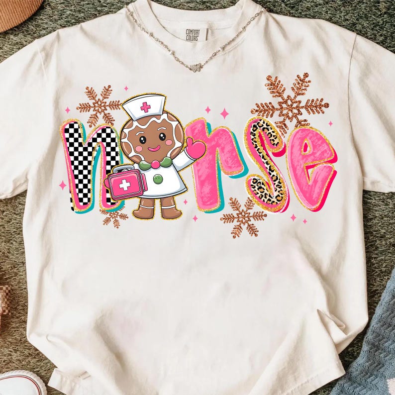May include: Cream-colored t-shirt with a colorful "Nurse" graphic. The "N" is checkered, the "R" and "S" are pink with leopard print accents, and the "U" and "E" are pink. A gingerbread nurse with a medical bag is in the center.