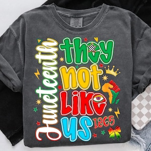 May include: A dark gray sweatshirt with the words "Juneteenth" and "They Not Like Us" in colorful text. The design includes stars, a crown, and the year 1865, celebrating the Juneteenth holiday.