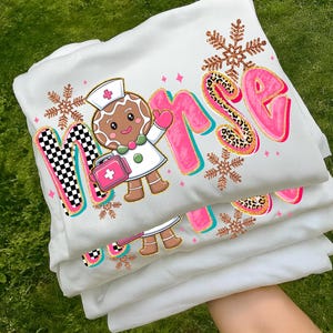 May include: White sweatshirt with a colorful "Nurse" graphic. The design features a gingerbread nurse, snowflakes, and the word "Nurse" in pink with leopard print accents. The sweatshirt is folded and stacked.