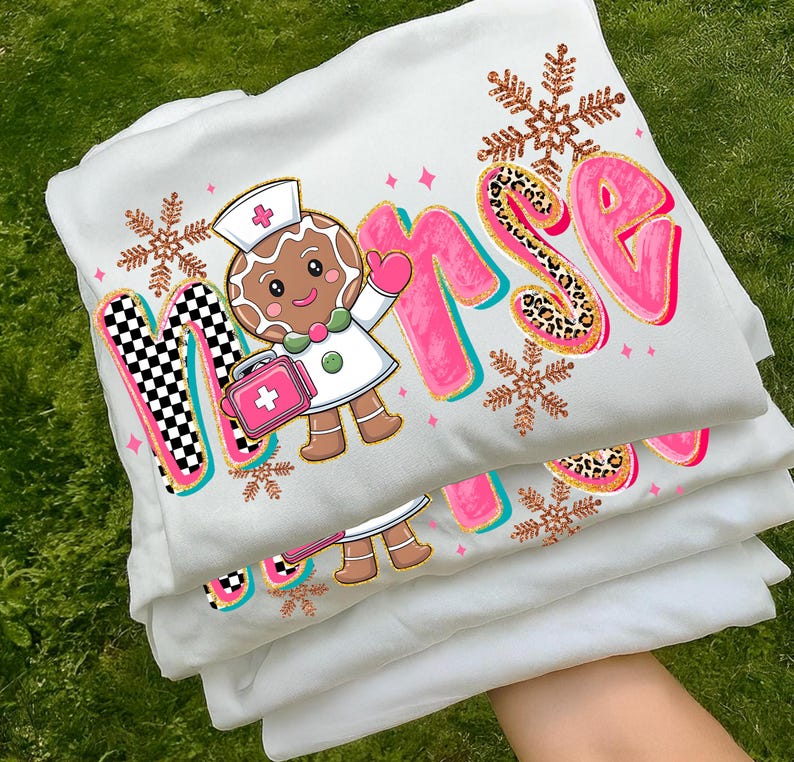 May include: White sweatshirt with a colorful "Nurse" graphic. The design features a gingerbread nurse, snowflakes, and the word "Nurse" in pink with leopard print accents. The sweatshirt is folded and stacked.